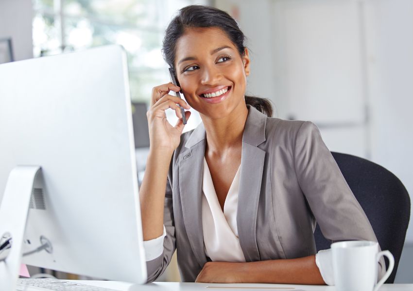 A smiling property manager with a grey suit jacket and long dark hair sits in front of a desk computer while discussing a rental property with their client.