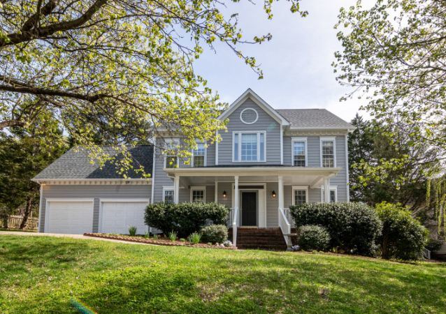 A grey pannelled Carrollton single family home, behind a well trimmed green lawn, is pictured under the shade of tree branches.