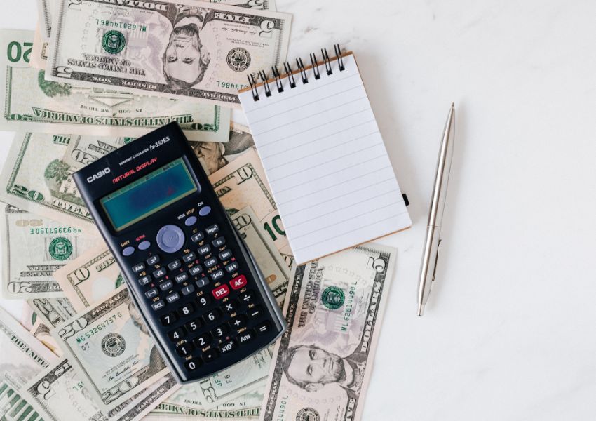 American bills, a small notepad, a silver pen and a black calculator sit on a white desk.