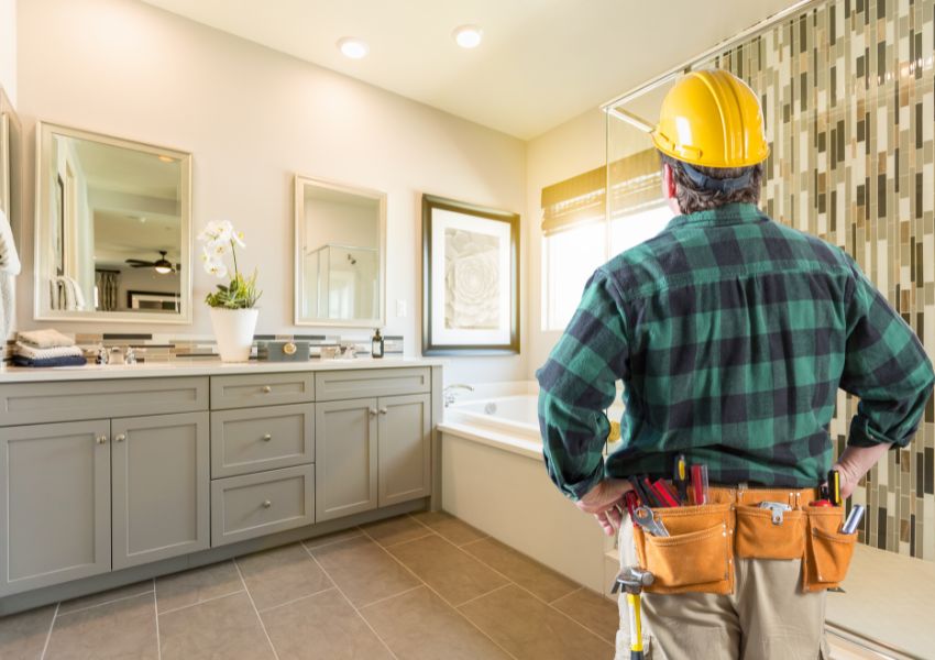 A contractor with a tool belt and yellow hard hat stands facing a neutral toned and renovated bathroom.