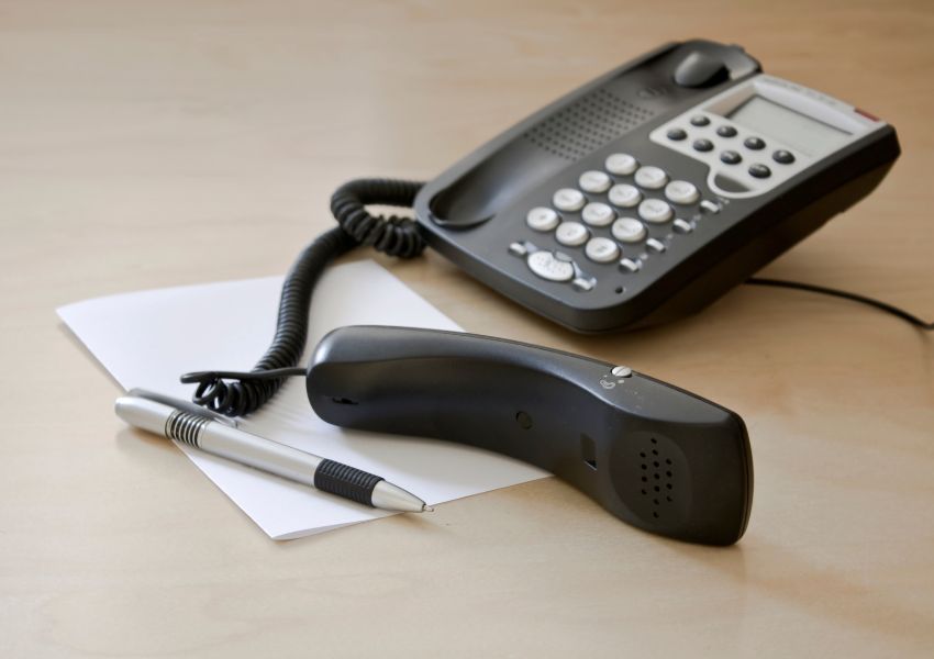 A black landline sits on a light wooden desk, with the phone lying on a piece of paper.