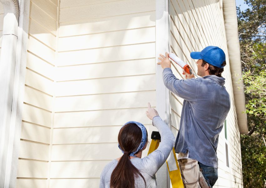 Two McKinney property managers work to maintain the exterior of an investment property by painting the trim of a cream panelled house white.