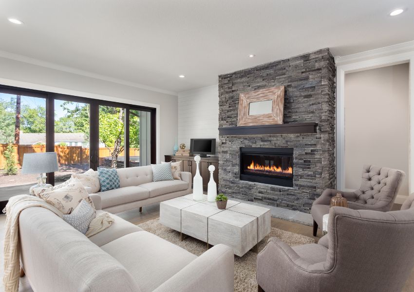 The interior of a cream living room with a dark gray brick fireplace is pictured, a marble coffee table and floor to ceiling windows on the back wall.