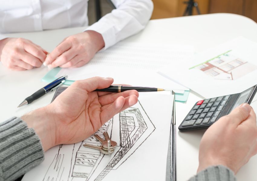 Two property managers price a rental property at a desk, as they calculate numbers on a grey calculator and examine the property's features using drawings.