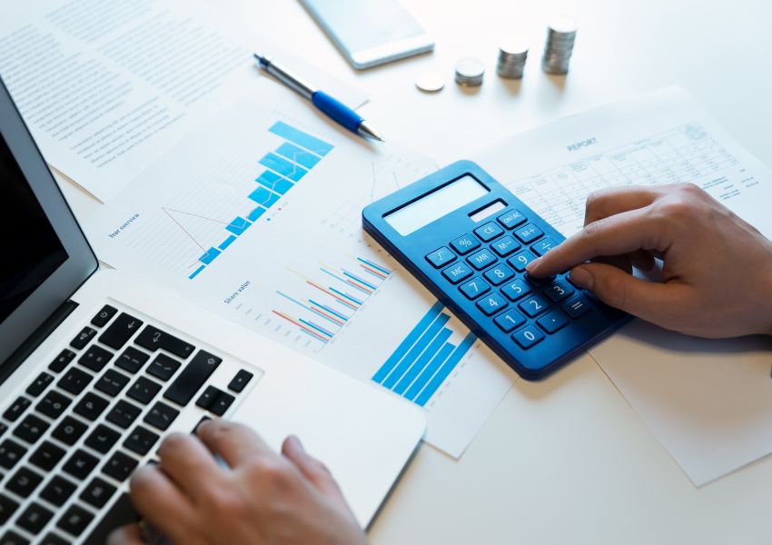 A landlord’s hands are pictured calculating their investment budget with a calculator, as well as market trend projections with figures and graphs in front of their hands.