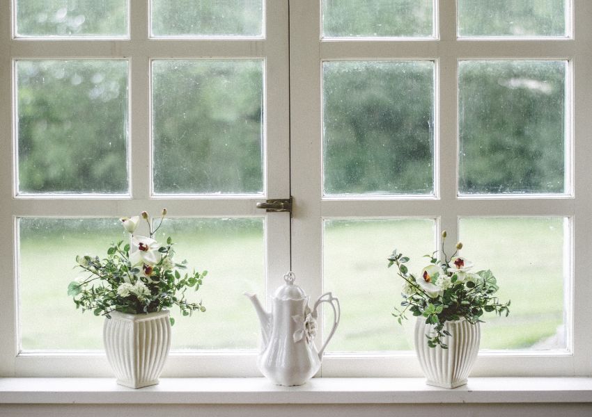 The inside of a white panelled window is pictured overlooking a green lawn, with two plants and a teapot on the windowsill.