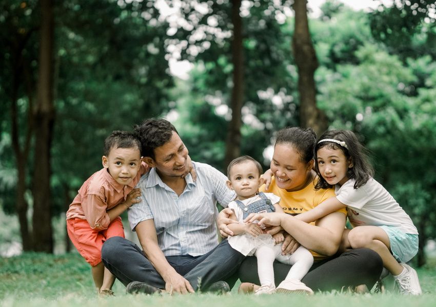A family of five sits on the grass of a park, with two parents hugging their three young children.